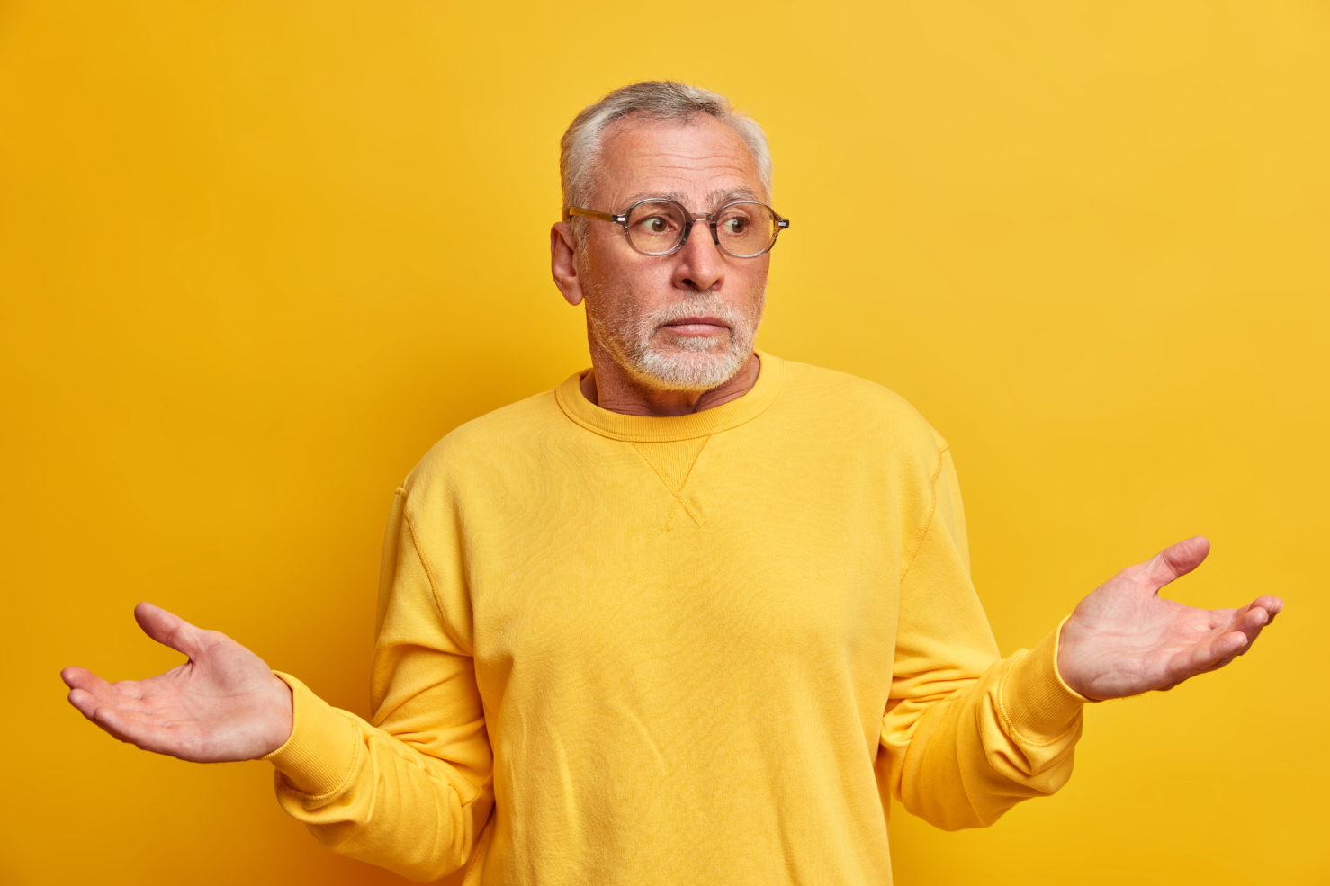 Portrait of puzzled hesitant bearded mature man spreads palms and faces difficult choice looks uncertain wears casual jumper optical glasses has confused expression isolated over yellow background